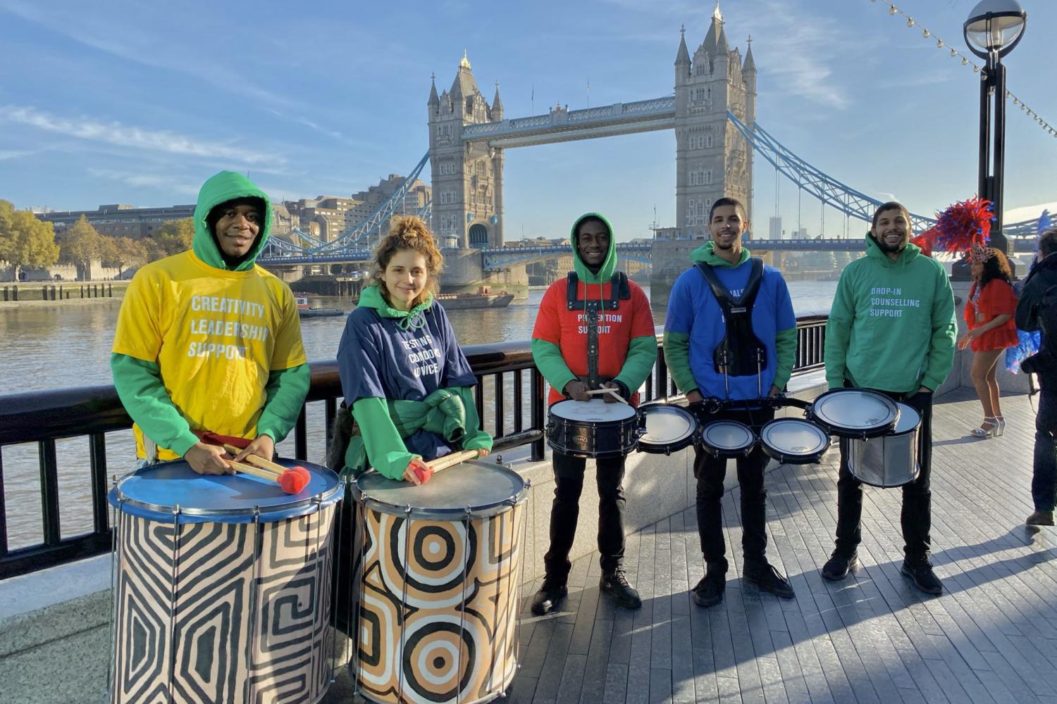 Group shot of smiling people wearing METRO hoodies, holding drums, with Tower Bridge in the background