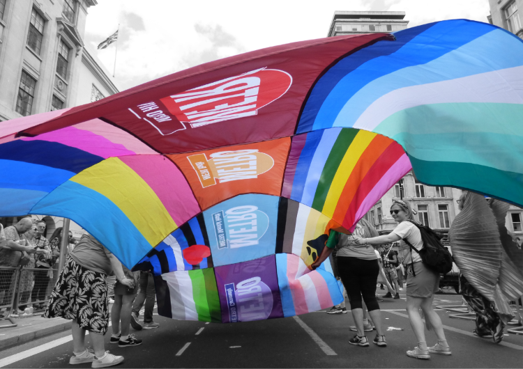  People proudly carrying a large rainbow-striped flag featuring multiple LGBTQ+ pride flags and METRO branding during a Pride march through central London.