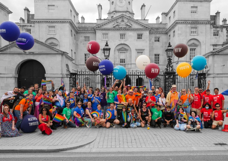 Large diverse group of METRO volunteers and supporters gathered in front of Horse Guards Parade, holding colourful balloons with "HIV" and other messages, celebrating Pride with rainbow flags and bright clothing.