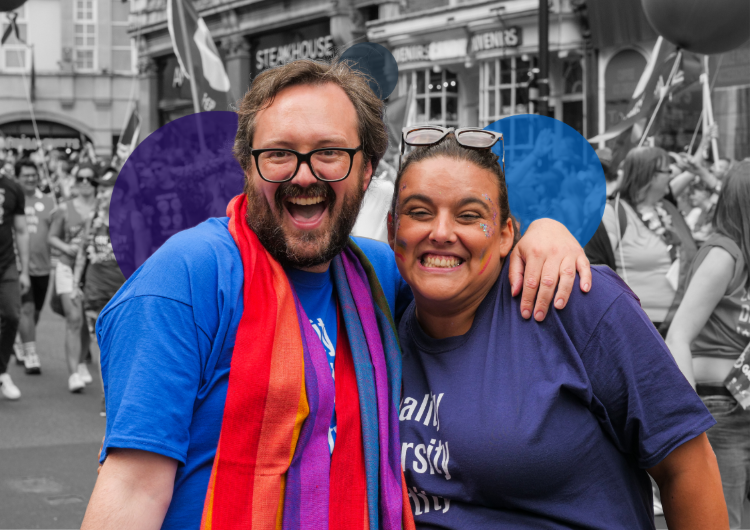 Two METRO volunteers sharing a joyful moment at Pride, one wearing rainbow scarf and glasses, both beaming with happiness in their purple METRO t-shirts.