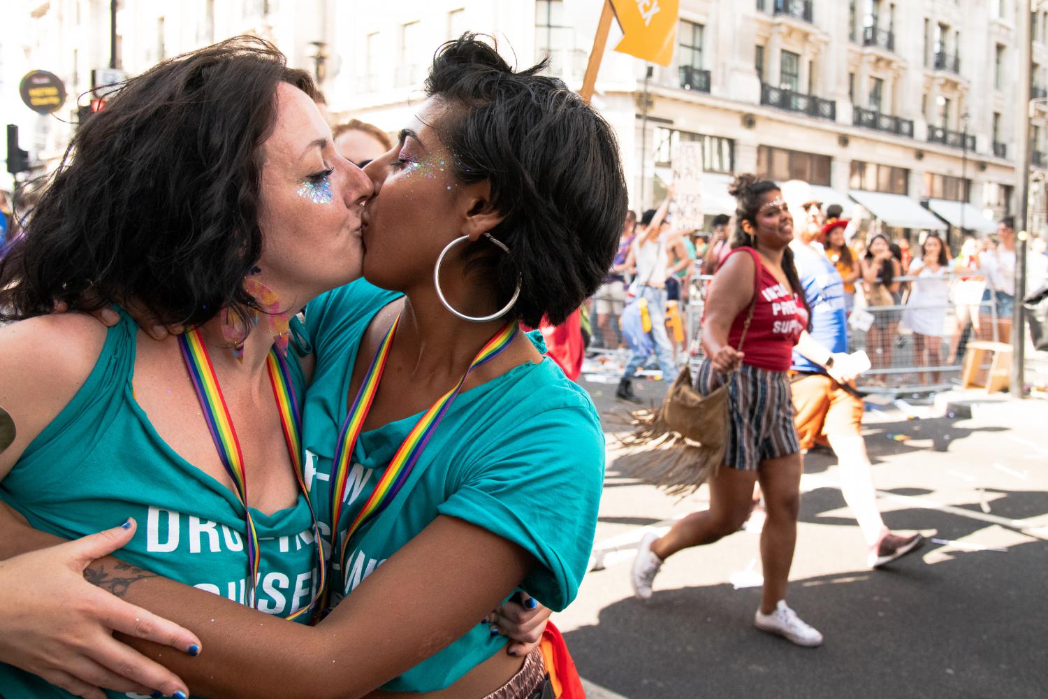 Photo of two women embracing and kissing at Pride. One is Black, one is white. They are both wearing METRO t-shirts and rainbow lanyards, and have glitter on their faces. 
