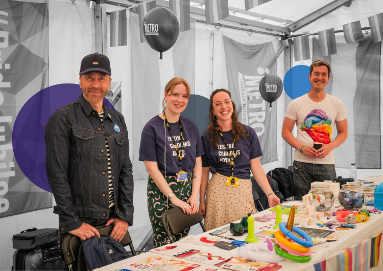 Four smiling METRO volunteers standing behind an information table at an event, with rainbow merchandise and leaflets displayed, ready to welcome visitors.