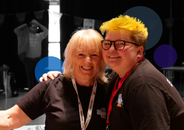 Older woman with blonde hair embracing a young person with bright yellow hair and glasses at a METRO event, both wearing lanyards and sharing a genuine, heartfelt moment.